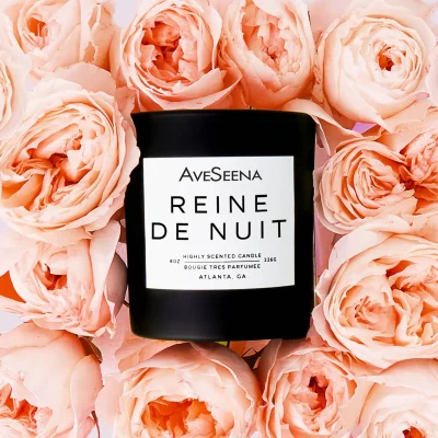 Reine de Nuit candle surrounded by pink roses, featuring a black container with white label