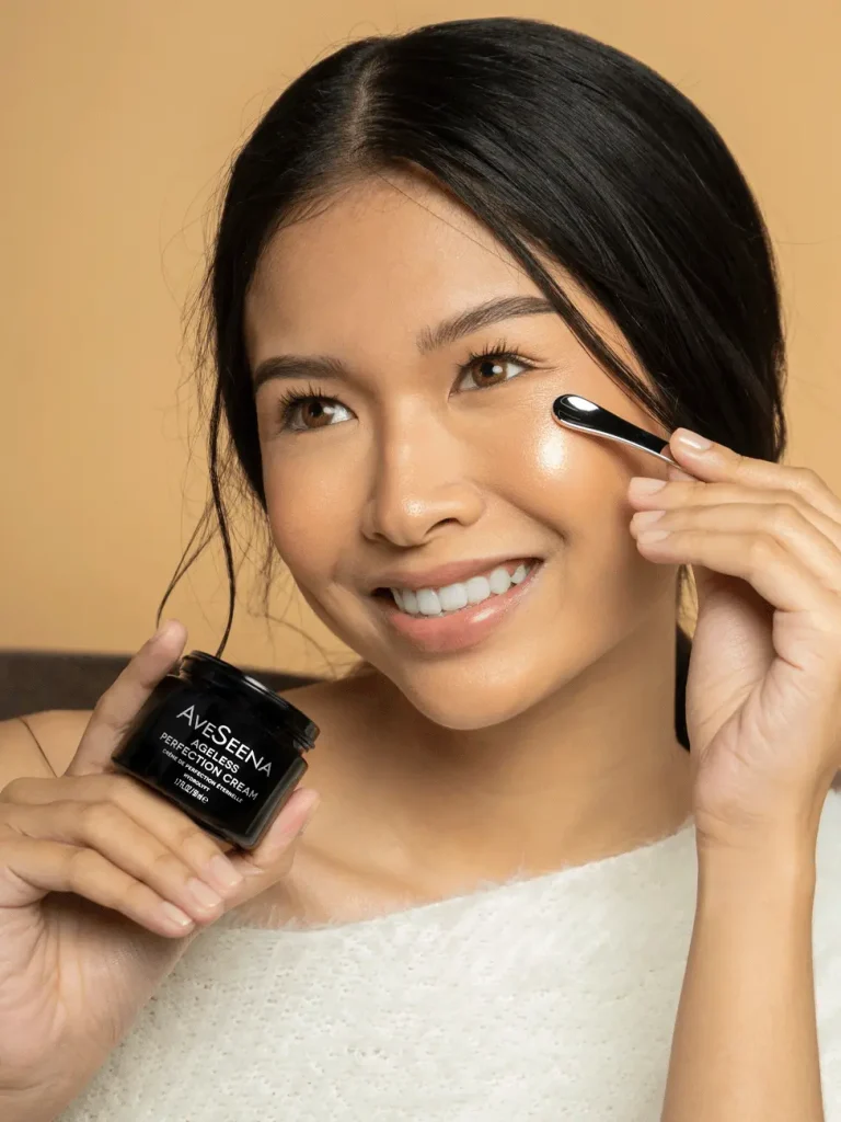 A woman smiling applies face cream from a jar labeled "Avesena" with a small spatula. She holds the jar near her face, radiating a joyful, fresh vibe.