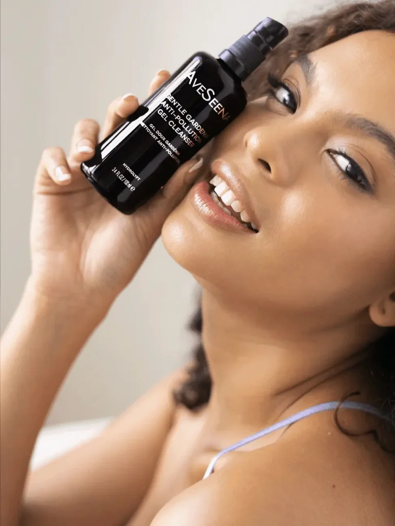 A woman smiles, holding a bottle of gel cleanser to her cheek. She exudes a fresh, radiant look. The product label reads "Gentle Gardenia Gel Cleanser."